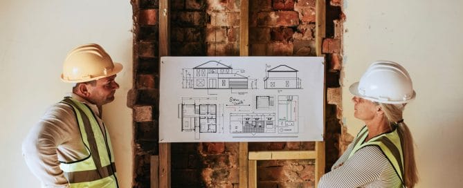 Two workers in high-visibility jackets and hard-hats looking at an architectural design in a room under construction.
