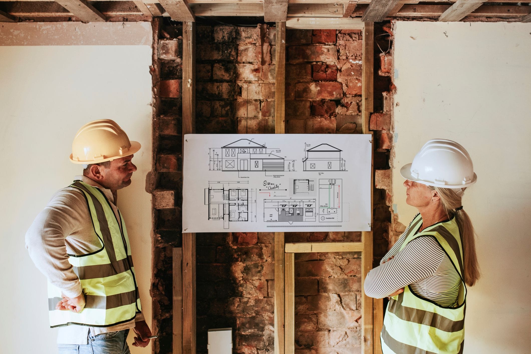 Two workers in high-visibility jackets and hard-hats looking at an architectural design in a room under construction.