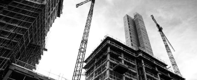 A black and white image of tall buildings and skyscrapers looking upwards from the ground. With two construction cranes. May of the buildings have scaffolding.